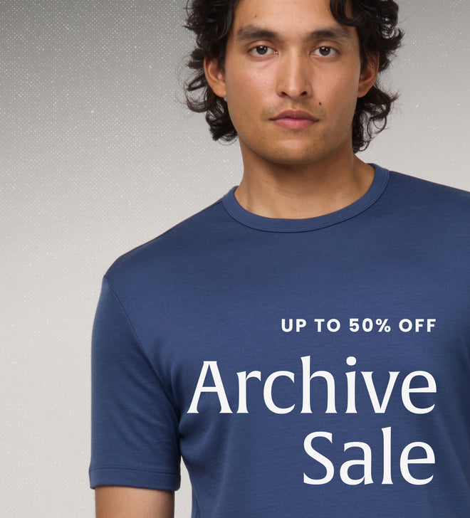 A man wearing a navy blue crew neck t-shirt set against a textured gray background, with white text overlay reading 'UP TO 50% OFF - Archive Sale'
